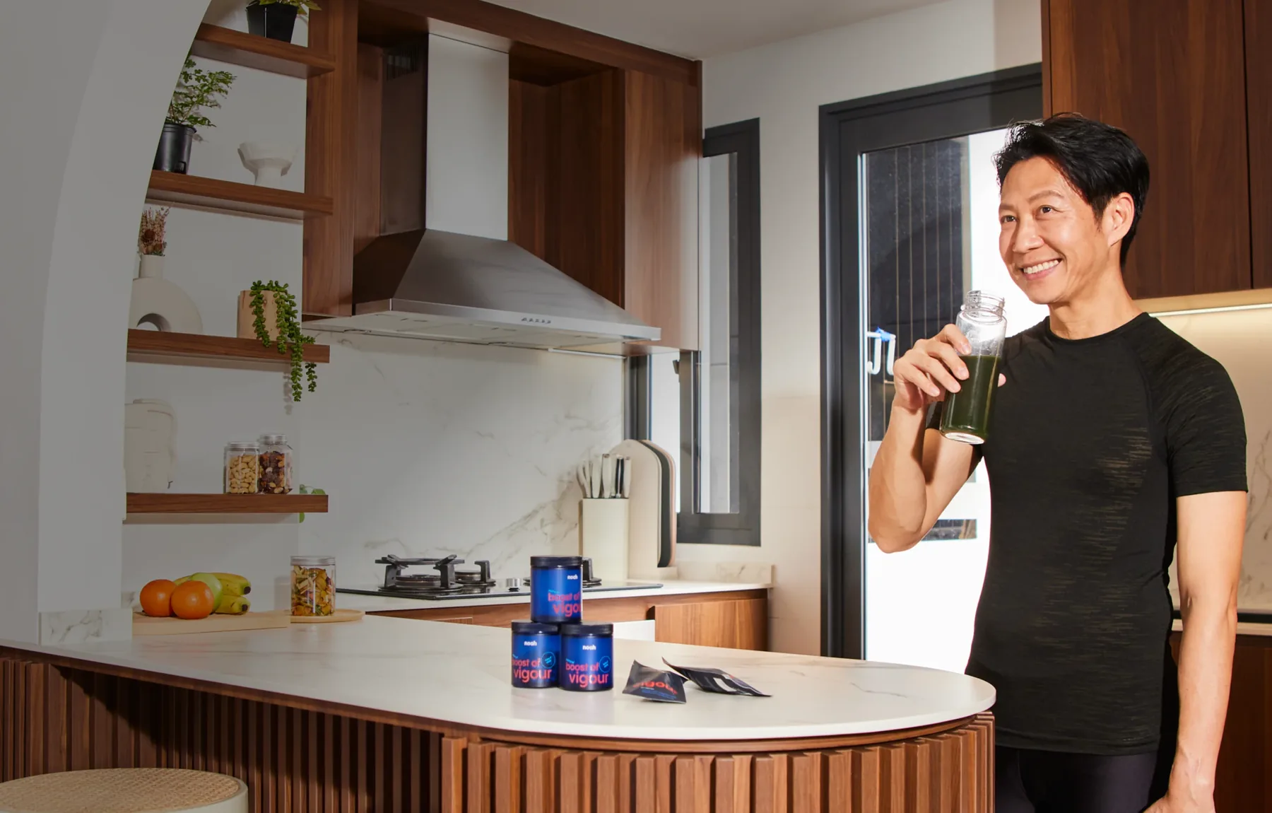 A man smiling in his kitchen at home, drinking a green juice, with Vigour on the counter
